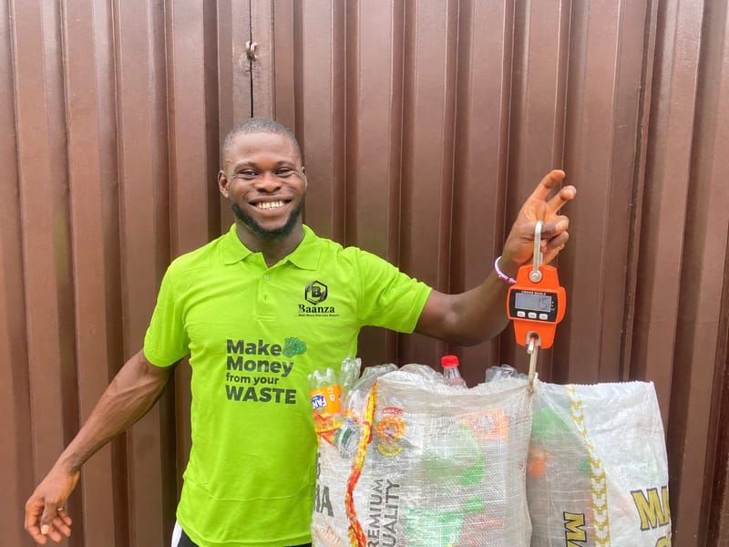 A smiling collector in a green Baanza shirt reading Make Money from your Waste holds up a hanging scale weighing a sack of plastic bottles.
